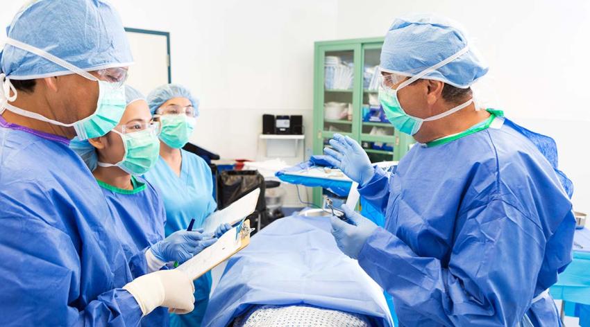 A male attending surgeon is teaching a group during an operationt in a hospital. They are reviewing patient's chart and prepping her for surgery. Surgeons are wearing sterile gowns, caps, masks, and gloves
