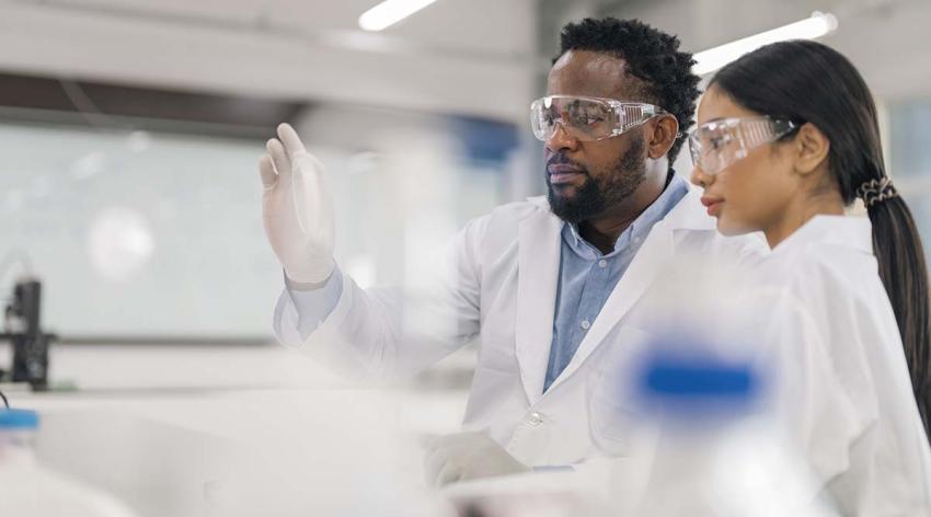 Two researchers wearing lab coats and eye protection analyze a sample in a laboratory. 