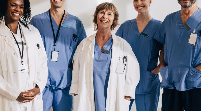 Five doctors, three in blue scrubs and two wearing white coats, smiling in front of a white wall