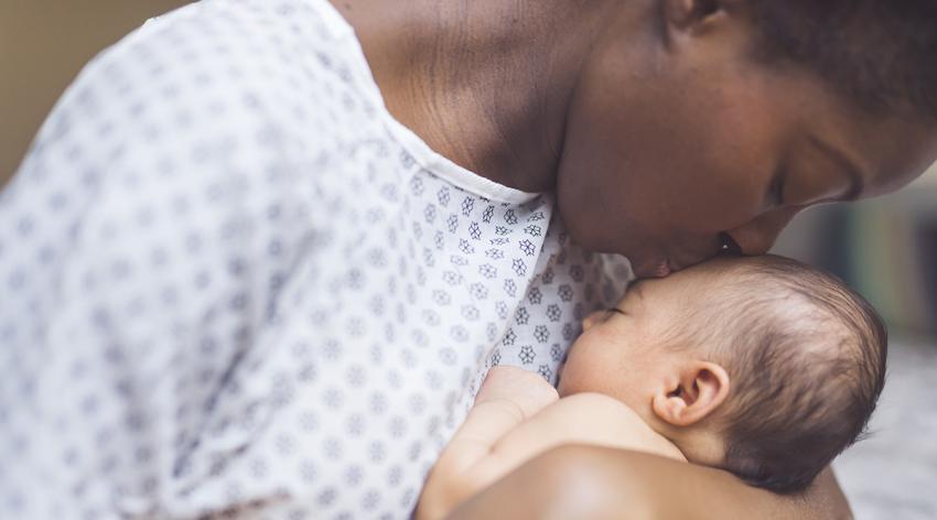 A young African American mother in a hospital gown gently holds her infant in her arms and smiles down at her. The baby's eyes are closed.