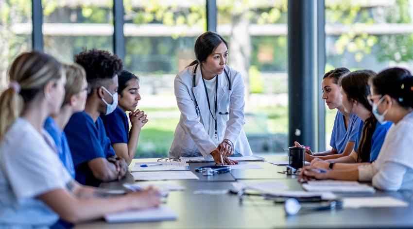 A small group of medical student residents gather around a boardroom table to to meet with their medical team lead. They each have cases out on front of them as they work together collaboratively to discuss each one. They are each dressed professionally in medical scrubs and are listening attentively to the doctor leading the meeting at the head of the table.