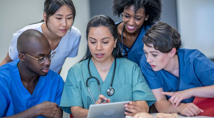 A group of medical students in training gather around to look at a tablet screen while studying.