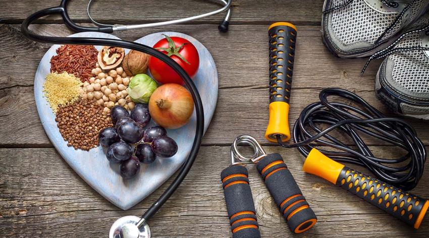 An overhead image showing grains, vegetables, and fruit on a heart-shaped white plate, while a stethoscope and some exercise equipment is also placed on the same wooden table with the food.  