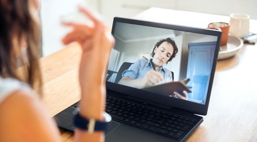 Psychotherapist seen on laptop screen writing on clipboard while listening to the female patient during an online counselling session.