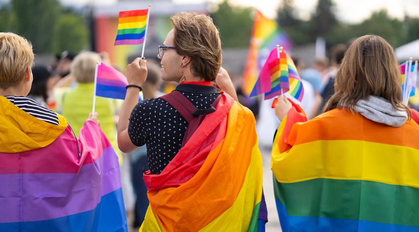 Wrapped in bisexual flag and pride flags, this trio are waving small pride flags and watching a gay pride event