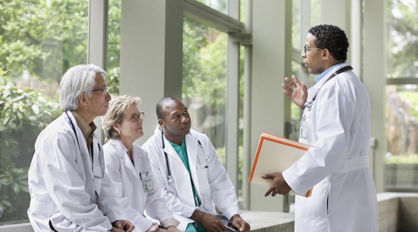 Three doctors in white coats sit on a bench while a fourth doctor stands and speaks