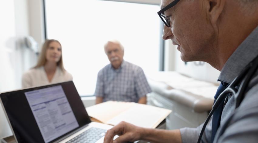 A doctor looks at an electronic medical record (EMR) on his laptop in the exam room with two patients in the background.