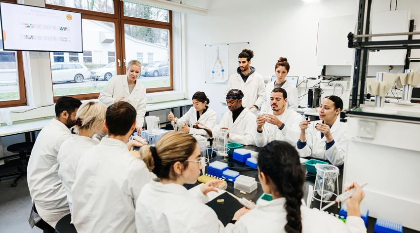 A group of students and a medical tutor working on a DNA experiment together in a laboratory around a large desk.