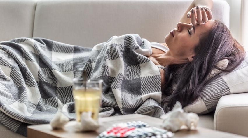 Woman having symptoms of Covid-19 lies covered in blanket in isolation with handkerchief and pills next to her.