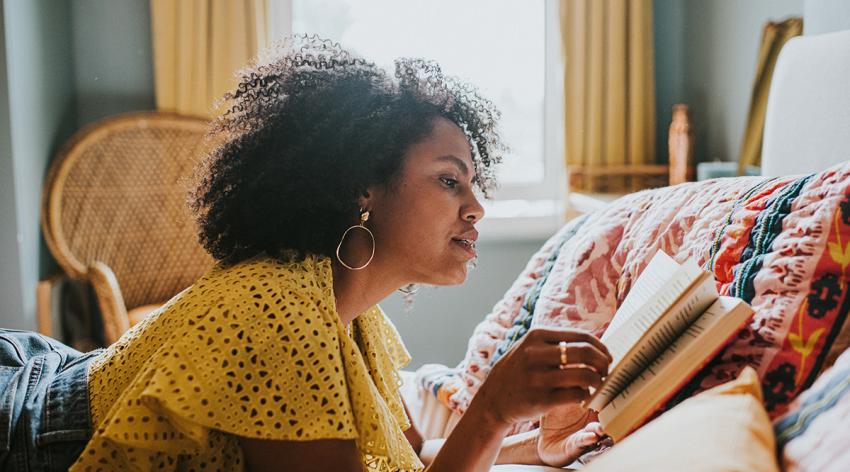 A woman lies on her stomach on a bed and reads a book