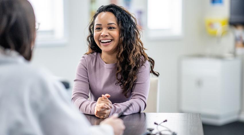 A young woman of mixed race, sits with her female doctor as they discuss her health concerns. The patient is dressed casually and has a smile on her face.
