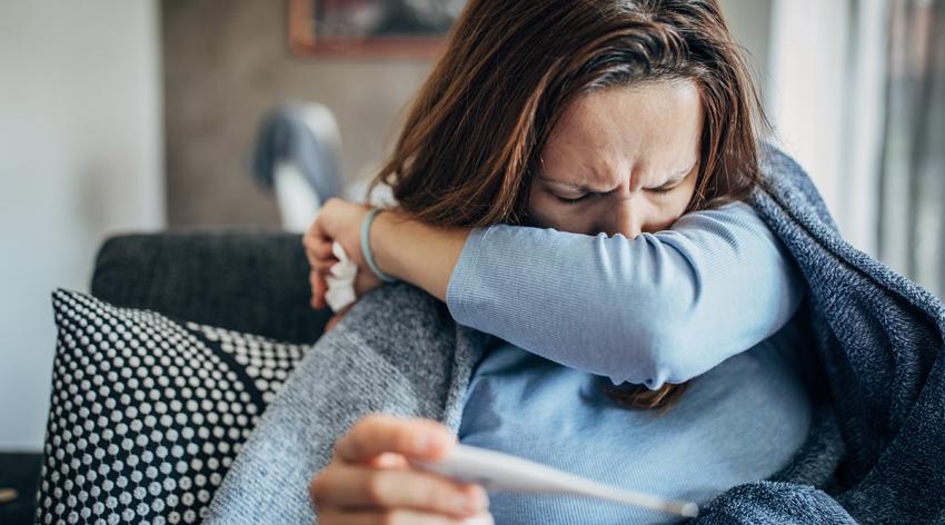 Woman with fever symptoms sitting on sofa and holding thermometer