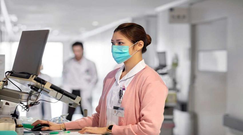 A woman doctor enters information into a computer while wearing a face mask.